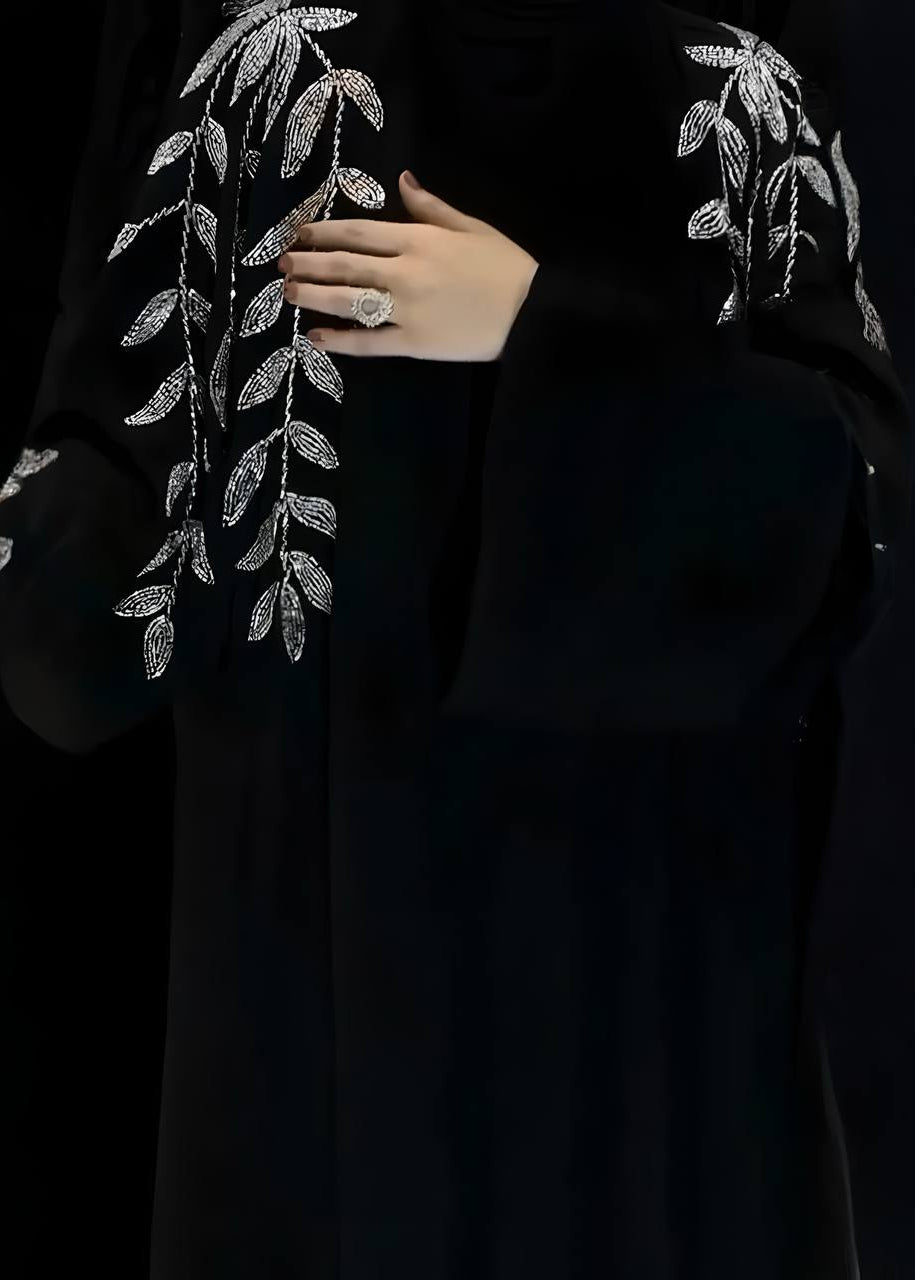 Black abaya with silver leaf embroidery held by a hand against a black background